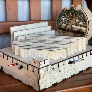 May include: A wooden cable organiser with a tree design and the words "Family is Everything". The organiser has slots for cords and a compartment for small items. The organiser is made of light wood and has a natural finish.