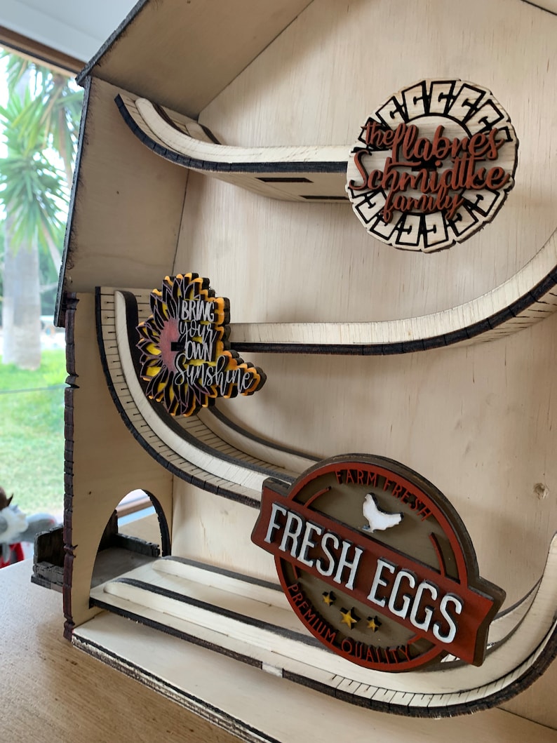 May include: A wooden farmhouse-style tiered shelf with three shelves. The top shelf has a brown circle with the text "The Labres Schmidtke Family". The middle shelf has a sunflower with the text "Bring Your Own Sunshine". The bottom shelf has a brown circle with the text "Farm Fresh Fresh Eggs Premium Quality".