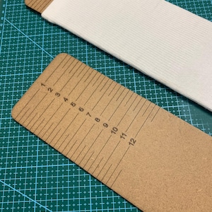 Puede incluir: Primer plano de dos reglas de madera con marcas en pulgadas, junto con una tela blanca. Las reglas están marcadas de 1 a 12 pulgadas. La tela blanca tiene una textura acanalada. El fondo es una esterilla de corte verde. Las reglas miden hasta 30,48 cm.