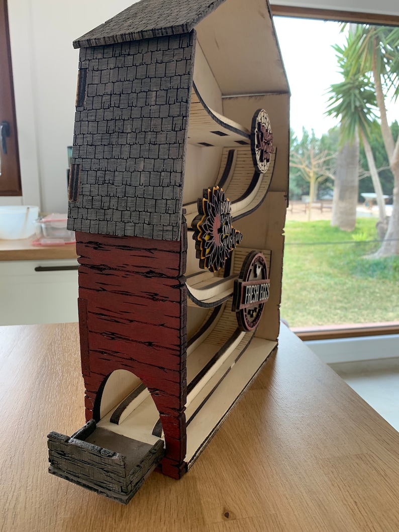 May include: A wooden model of a barn with a red and gray exterior. The barn has a series of ramps and wheels inside, with signs that read "Fresh Eggs" and "Brown Eggs From Our Chicks".