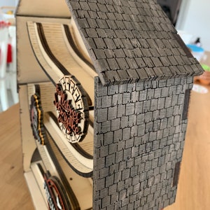 May include: A wooden farmhouse-style egg holder with a gray shingled roof and a sign that reads "Fresh Eggs". The holder has a wooden ramp for the eggs to roll down.