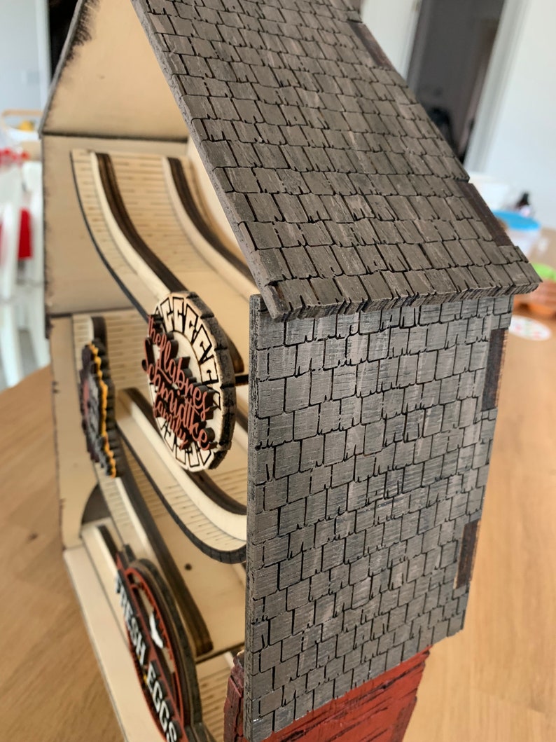May include: A wooden farmhouse-style egg holder with a gray shingled roof and a sign that reads "Fresh Eggs". The holder has a wooden ramp for the eggs to roll down.