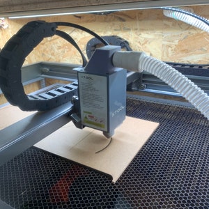 May include: A close-up of a laser cutter engraving a piece of wood. The laser cutter is a gray and black machine with a clear hose attached to it. The machine is sitting on a black metal grid.