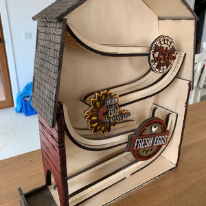 May include: A wooden farmhouse-style structure with a red brick facade and a gray shingled roof. The structure has three levels with a curved track on each level. The track is decorated with a sunflower, a sign that says "Bring on the Sunshine", and a sign that says "Fresh Eggs".