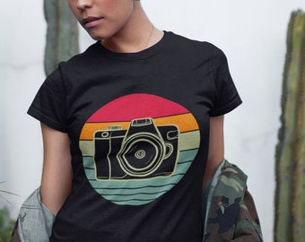 May include: Black t-shirt featuring a retro camera design within a circular graphic with red, orange, yellow, green, and blue stripes. The model is wearing a camouflage jacket and denim shorts. The background includes cacti.