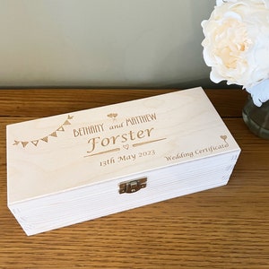 May include: A white wooden box with a hinged lid and a metal clasp. The box is engraved with the text "Bethany and Matthew Forster 13th May 2023 Wedding Certificate" and a design of bunting and balloons.