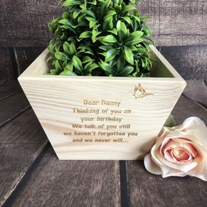 May include: A wooden planter box with a personalised message engraved on the front. The message reads "Dear Danny Thinking of you on your birthday We talk of you still we haven't forgotten you and we never will..." The planter is filled with a green plant and a pink rose is sitting next to it.
