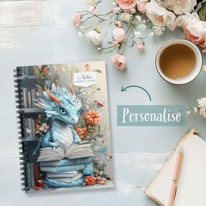 Può includere: Diario di lettura a spirale con un'illustrazione di un drago blu che legge libri, circondato da fiori. Il diario presenta il testo "Nesta's Reading Journal" e la parola "Personalise".