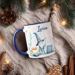 Puede incluir: Taza de cerámica blanca con asa y borde azules, con una ilustración de mago y el nombre "Lucian". El mago lleva un sombrero azul y un bastón, cerca de un portátil. La taza está decorada con estrellas y elementos florales.