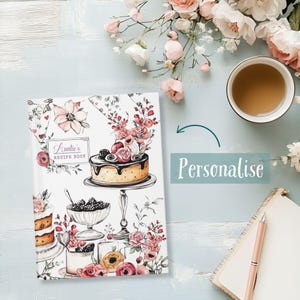 Puede incluir: Un libro de recetas titulado "Auntie's Recipe Book" con ilustraciones florales y la palabra "Personalise". La portada presenta un pastel y otros postres. También se ven una taza de té, flores, un cuaderno y un bolígrafo.