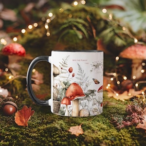 Puede incluir: Taza de cerámica blanca con asa y borde negros, con un diseño forestal caprichoso con setas, helechos y el texto "Into the Forest I Go". La taza está colocada sobre un fondo cubierto de musgo con follaje otoñal.