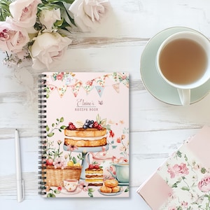 Può includere: Un ricettario rilegato a spirale con copertina rosa, con un'illustrazione ad acquerello di una torta, un servizio da tè ed elementi floreali. La copertina recita "Elaine's Recipe Book". Sono visibili anche una penna e una tazza di tè.