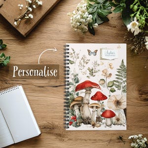Può includere: Un taccuino a spirale con un'illustrazione ad acquerello di funghi e piante. La copertina presenta funghi rossi e marroni, felci e farfalle. Il testo "Sophia's Field Notes" è in alto a destra. La parola "Personalizza" è anche visibile.