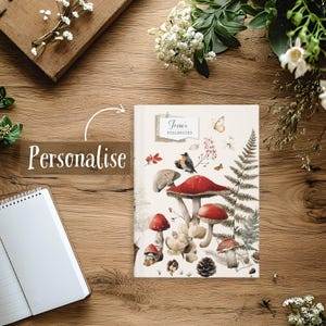 May include: A personalized notebook with a mushroom and forest illustration. The cover features red-capped mushrooms, a bird, and ferns. The text "Irene's Fieldnotes" is printed on a label. The notebook is on a wooden surface with floral accents.