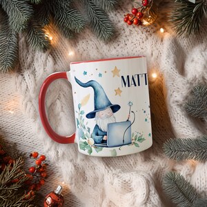 May include: A white ceramic mug with a red handle and rim. The mug features a whimsical illustration of a wizard with a laptop, the name "MATT", and star accents. The mug is surrounded by festive holiday decorations.