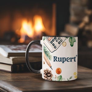 以下が含まれることがあります： 黒いハンドルと縁が付いた白いセラミックマグ。電卓、ペン、松ぼっくり、コインの水彩デザインが特徴です。「Rupert」という名前は濃い青色で印刷されています。マグカップは木製の表面に置かれています。