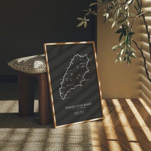 Może przedstawiać: Czarno-biały wydruk mapy szlaku Tour du Mont Blanc, oprawiony w naturalną drewnianą ramę. Mapa przedstawia trasę szlaku o długości 170 km, kt&oacute;ry okrąża masyw Mont Blanc w Alpach. Wydruk wisi na ścianie w pokoju z drewniana podłogą.