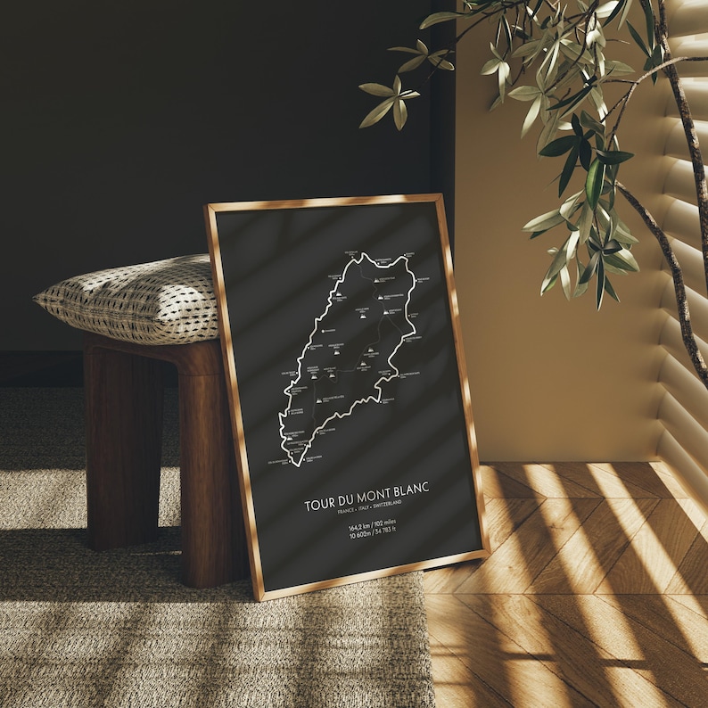 Może przedstawiać: Czarno-biały wydruk mapy szlaku Tour du Mont Blanc, oprawiony w naturalną drewnianą ramę. Mapa przedstawia trasę szlaku o długości 170 km, kt&oacute;ry okrąża masyw Mont Blanc w Alpach. Wydruk wisi na ścianie w pokoju z drewniana podłogą.