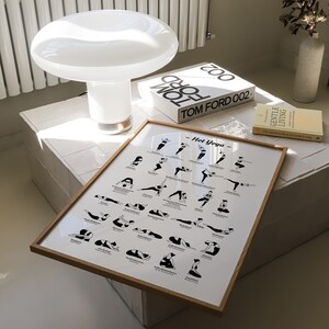May include: A framed poster with a white background and black illustrations of yoga poses. The poster is titled "Hot Yoga" and includes the names of each pose. The poster is sitting on a white tiled surface with a white lamp and a book titled "TOM FORD 002" in the background.
