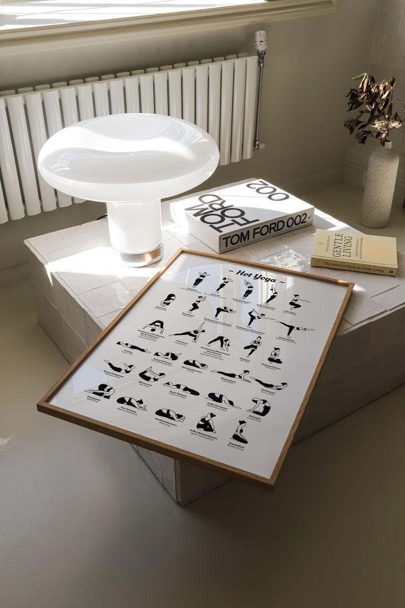 May include: A framed poster with a white background and black illustrations of yoga poses. The poster is titled "Hot Yoga" and includes the names of each pose. The poster is sitting on a white tiled surface with a white lamp and a book titled "TOM FORD 002" in the background.