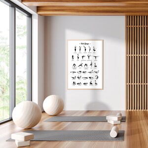 May include: A white framed poster with the title "Hot Yoga" and a black and white illustration of yoga poses. The poster is hanging on a white wall in a room with a wooden floor, two yoga mats, and a yoga ball.