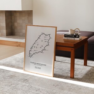 Może przedstawiać: Czarno-biały wydruk mapy szlaku Tour du Mont Blanc w drewnianej ramie. Mapa przedstawia trasę szlaku, zmiany wysokości i kluczowe punkty. Wydruk jest wyświetlany na drewnianym stole z książką i ceramicznym kubkiem.