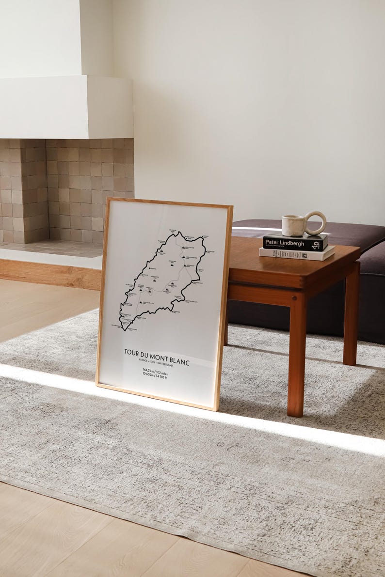 Może przedstawiać: Czarno-biały wydruk mapy szlaku Tour du Mont Blanc w drewnianej ramie. Mapa przedstawia trasę szlaku, zmiany wysokości i kluczowe punkty. Wydruk jest wyświetlany na drewnianym stole z książką i ceramicznym kubkiem.