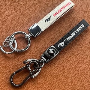 Peut inclure: Deux porte-clés en cuir noir et blanc avec des anneaux et des clips en métal argenté. Les deux porte-clés présentent le mot "MUSTANG" en lettres rouges et un logo de cheval stylisé.