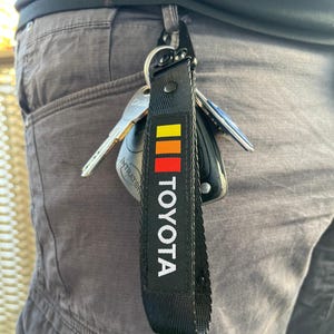 Könnte beinhalten: Schwarzer Schlüsselanhänger mit einem schwarz-weißen "TOYOTA"-Logo und einem roten, orangefarbenen und gelben Streifenmuster.