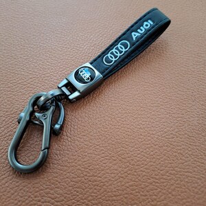 K&ouml;nnte beinhalten: Schwarzer Leder-Schl&uuml;sselanh&auml;nger mit einem silbernen Metallverschluss und einem silbernen Audi-Logo aus Metall. Der Schl&uuml;sselanh&auml;nger tr&auml;gt den Schriftzug "Audi" in silberner Pr&auml;gung.