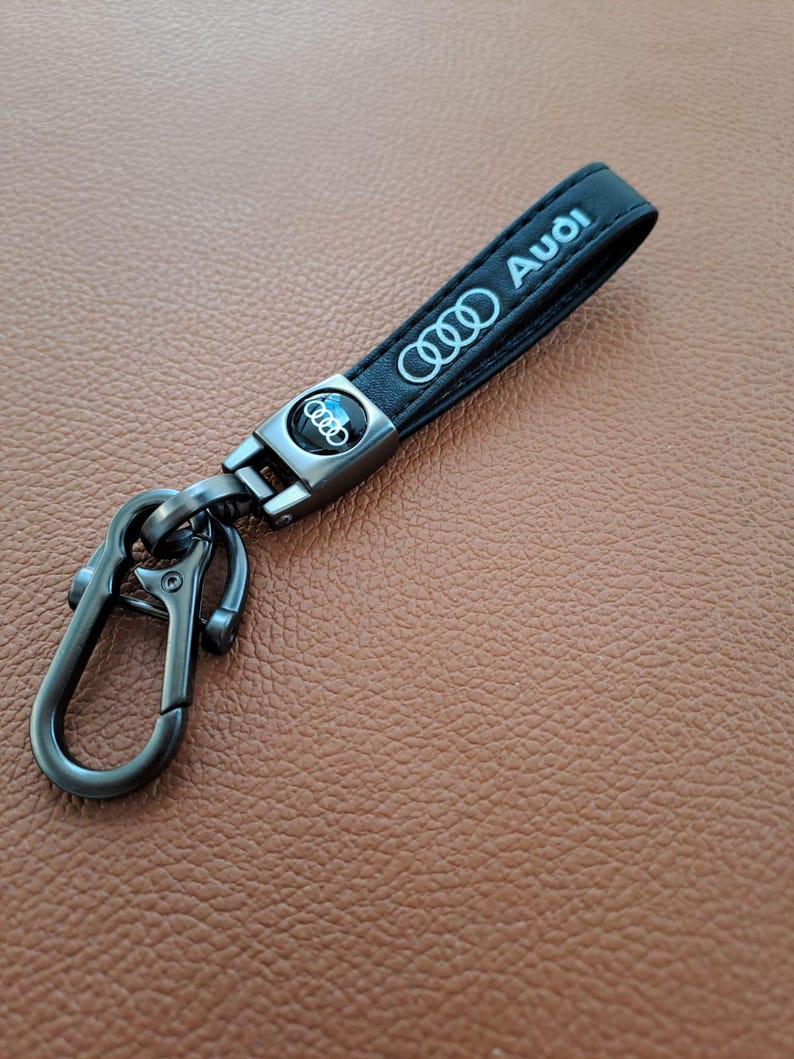 K&ouml;nnte beinhalten: Schwarzer Leder-Schl&uuml;sselanh&auml;nger mit einem silbernen Metallverschluss und einem silbernen Audi-Logo aus Metall. Der Schl&uuml;sselanh&auml;nger tr&auml;gt den Schriftzug "Audi" in silberner Pr&auml;gung.