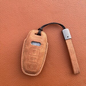 K&ouml;nnte beinhalten: Ein brauner Leder-Schl&uuml;sselanh&auml;nger mit einem schwarzen Band und einem silbernen Schl&uuml;sselring. Der Anh&auml;nger hat vier Kn&ouml;pfe und ein kleines Fenster.