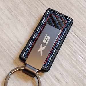 May include: A rectangular black leather keychain with a silver metal plate featuring the embossed text "X5". The keychain has a carbon fiber pattern and is accented with red, white, and blue stitching. A silver key ring is attached.
