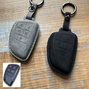 May include: Two leather key fob covers, one gray and one black, with silver key rings. The covers have embossed buttons and are stitched around the edges. The key fobs are on a wooden surface.