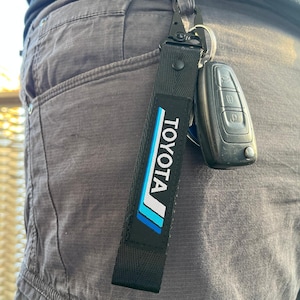 Pode incluir: Chaveiro preto com um logotipo Toyota azul e branco. O chaveiro está preso a um conjunto de chaves.
