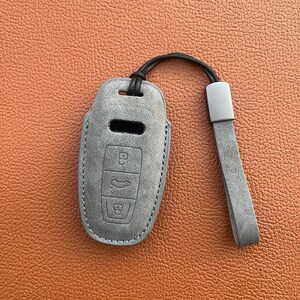K&ouml;nnte beinhalten: Ein grauer Leder-Schl&uuml;sselanh&auml;nger mit einem schwarzen Band. Der Anh&auml;nger hat drei Kn&ouml;pfe, einen mit einem Schlosssymbol, einen mit einem Autosymbol und einen mit einem Entriegelungssymbol.