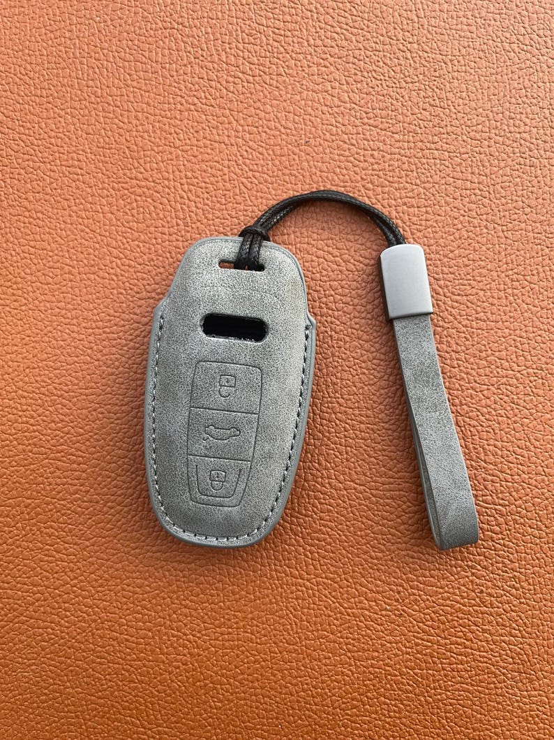 K&ouml;nnte beinhalten: Ein grauer Leder-Schl&uuml;sselanh&auml;nger mit einem schwarzen Band. Der Anh&auml;nger hat drei Kn&ouml;pfe, einen mit einem Schlosssymbol, einen mit einem Autosymbol und einen mit einem Entriegelungssymbol.