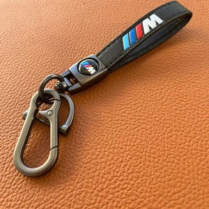 May include: Black leather keychain with a silver metal loop and a blue, red, and white M logo.