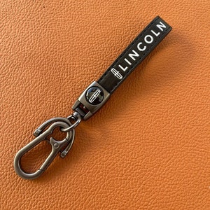 May include: A black leather keychain with a silver metal clasp and a silver metal Lincoln logo. The keychain has the word "LINCOLN" printed in white on the leather strap.