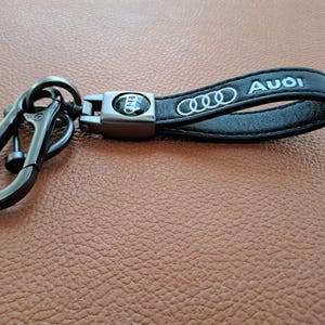 K&ouml;nnte beinhalten: Schwarzer Leder-Schl&uuml;sselanh&auml;nger mit einem silbernen Metallring und einem silbernen Audi-Logo aus Metall. Der Schl&uuml;sselanh&auml;nger tr&auml;gt den Schriftzug "Audi" in silberner Pr&auml;gung.