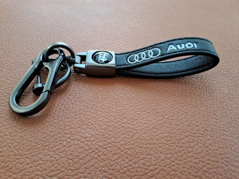 K&ouml;nnte beinhalten: Schwarzer Leder-Schl&uuml;sselanh&auml;nger mit einem silbernen Metallring und einem silbernen Audi-Logo aus Metall. Der Schl&uuml;sselanh&auml;nger tr&auml;gt den Schriftzug "Audi" in silberner Pr&auml;gung.