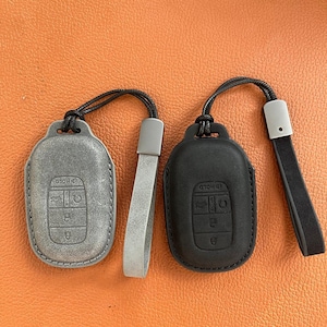 Peut inclure: Deux étuis de porte-clés en cuir, un gris et un noir, avec des lanières assorties. Le couvercle gris a un aspect vieilli, tandis que le noir est lisse. Les deux ont des symboles de clés en relief et le mot "HOLD".