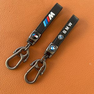 May include: Two black leather keychains with metal key rings. One keychain has a blue, red, and white BMW M logo. The other keychain has a black and white BMW logo.