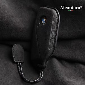 May include: Black leather key fob with a BMW logo and buttons. The fob has a black cord attached to it.