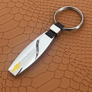 Peut inclure: Un porte-clés argenté avec une lanière en cuir noir et un charme argenté avec le mot "CHEVROLET" et un logo Chevrolet jaune.