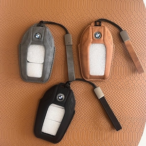 May include: Three leather keyrings with a BMW logo. The keyrings are in black, brown, and grey. Each keyring has a loop for attaching keys.