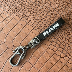 Può includere: Portachiavi in pelle nera con la scritta "RAM" a lettere bianche. Il portachiavi ha un moschettone color argento e un piccolo emblema circolare con "RAM". Lo sfondo è una superficie marrone strutturata.