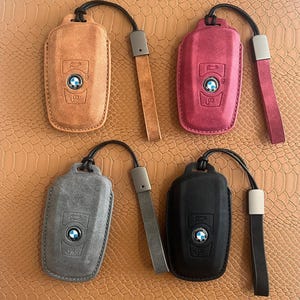 Peut inclure: Quatre housses en cuir pour clé de voiture BMW. Les housses sont en marron, rouge, gris et noir. Chaque housse est munie d'une sangle noire avec une boucle métallique.