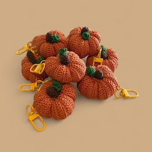 May include: A collection of orange crochet pumpkin keychains. Each pumpkin has a green leaf and brown stem detail. The keychains have yellow metal clasps. The pumpkins are arranged on a light brown surface.
