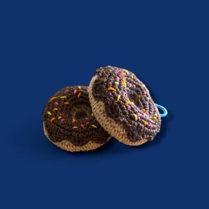 May include: Two handmade crochet donuts with chocolate frosting and colorful sprinkles. The donuts have a tan base and a dark brown frosting. One donut has a blue loop attached. The background is a solid dark blue.